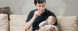 A young father holds a baby while smoking a cigarette.