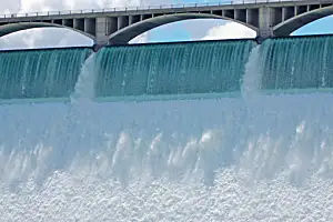 Water cascades over a dam, above which an arched bridge spans against a cloudy sky backdrop.