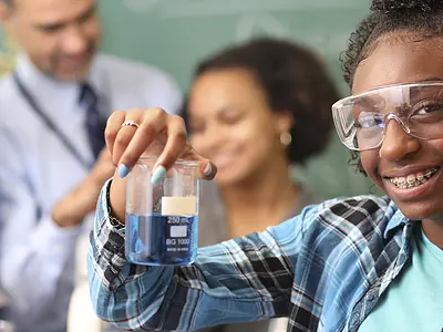 A teenage African American girl holds a flask in a science classroom while classmates and a teacher work in the background.