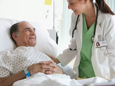 A female doctor stands next to the hospital bed of an older male patient, who is smiling.