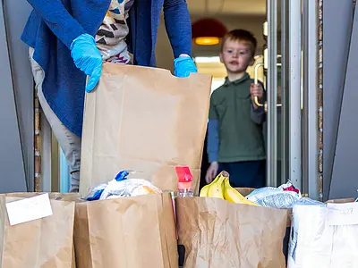 A delivery person brings bags of groceries to a family's front steps, while a boy watches from inside the door.