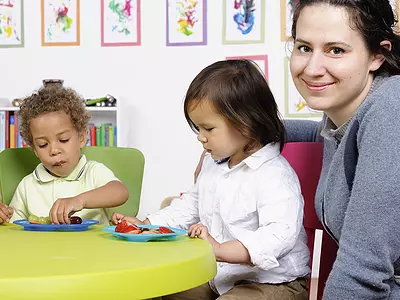 Three preschoolers eat fruit at a table with a teacher.