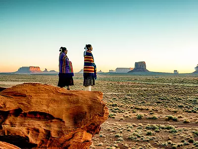 Two Navajo sisters in traditional clothing watch a sunset in Monument Valley.