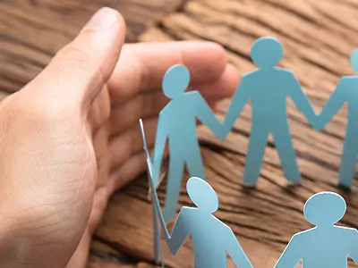 A paper cutout of people holding hands with human hands surrounding it.