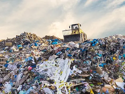 A bulldozer sits atop a heap of garbage in a large landfill.
