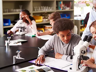 Two high school students use a microscope with help from a male science teacher.