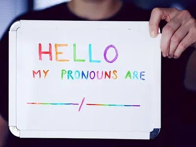 Photo of someone holding a whiteboard containing the words "Hello, my pronouns are" in rainbow colors