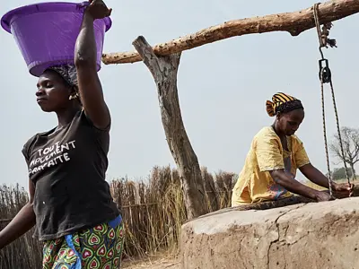 Photo of two African women working outside near a well. One is carrying a purple bucket on her head.