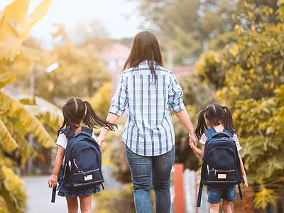 Photo of a mother holding hands with her two children wearing backpacks, walking away from the camera