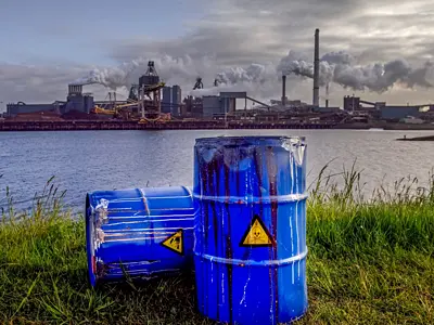 Photo of two barrels filled with hazardous waste on the banks of a river with a factory in the background
