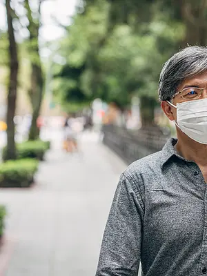 An Asian senior man wears a face mask and walks along a tree-lined sidewalk.