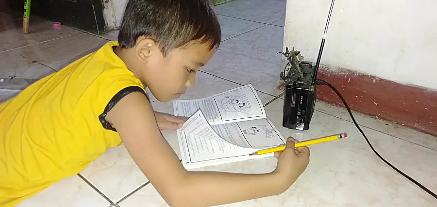 Child reading a book, holding a pencil on a tiled floor near a black radio.