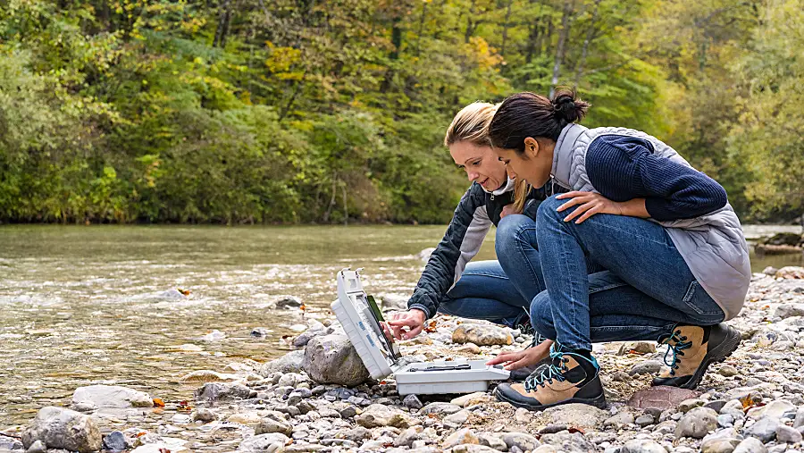 Two female scientists look at data on a tablet next to a creek.