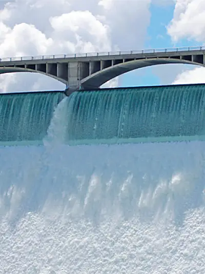 Water cascades over a dam, above which an arched bridge spans against a cloudy sky backdrop.