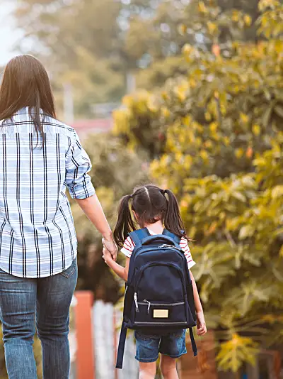 Photo of a mother holding hands with her two children wearing backpacks, walking away from the camera