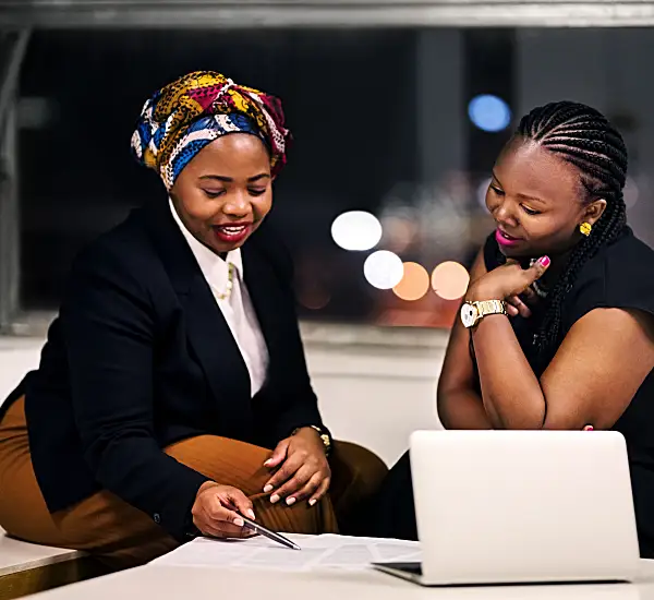 Two women review documents together, smiling, with city lights visible through a window.