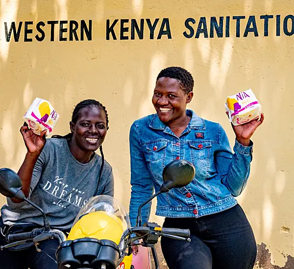 Photo of two Kenyan women smiling holding sanitary products near a motorbike