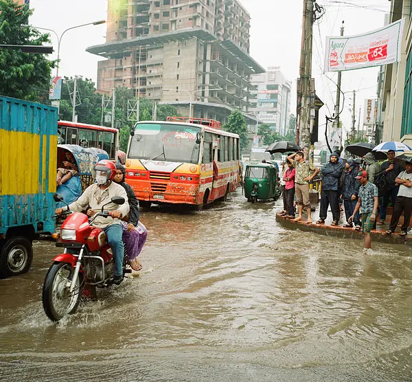 Flooding in Dhaka. People standing on the sidewalk. A motorcycle riding through the flooding.
