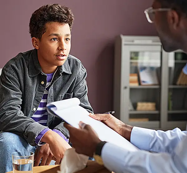 A clinician takes notes while talking with a young teenage boy.