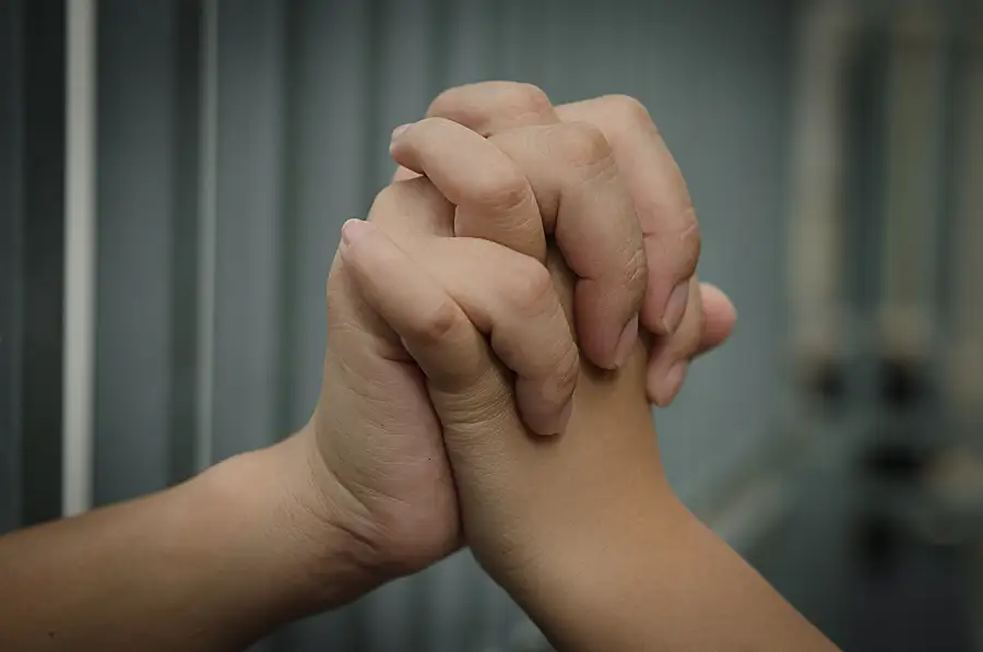 Close up photo of two linked hands, one reaching from behind bars in a correctional facility