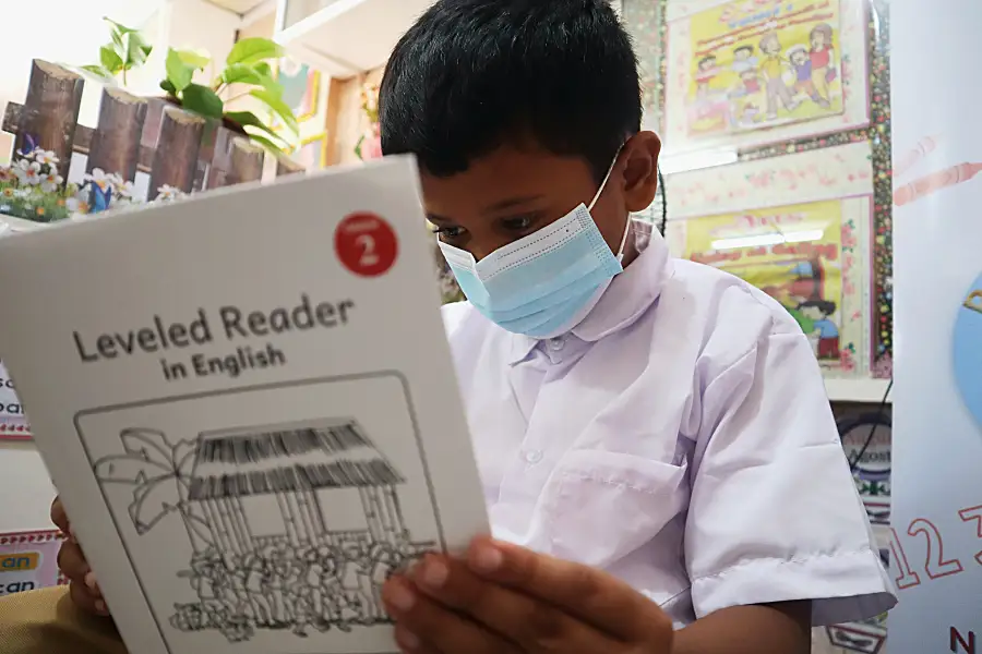 Child with a mask reads a book in English in a classroom