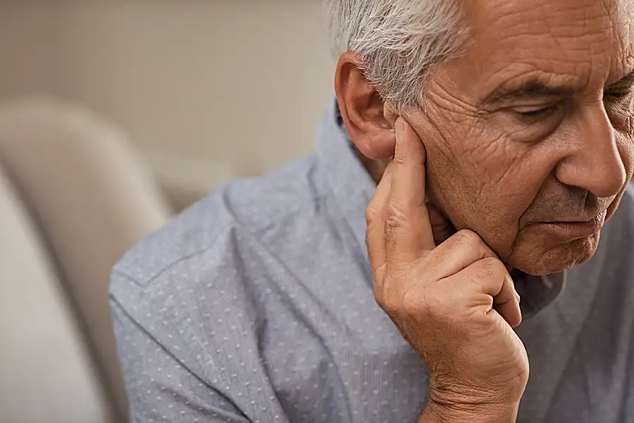 A senior white man looks down and points to his ear, as if he is having trouble hearing.