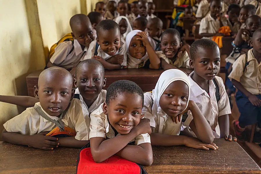 African schoolchildren crowd onto benches in a classroom and smile for the camera.