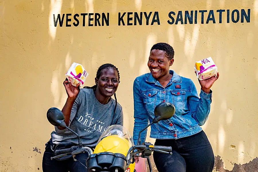 Photo of two Kenyan women smiling holding sanitary products near a motorbike