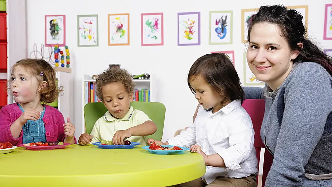 Three preschoolers eat fruit at a table with a teacher.