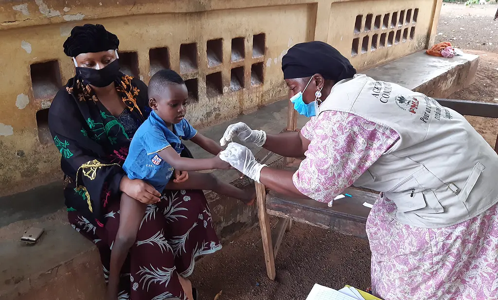 A community health worker performs a malaria rapid diagnostic test (RDT) on a child during a home visit in Guinea, while wearing a mask to adhere to COVID-19 safety guidelines.