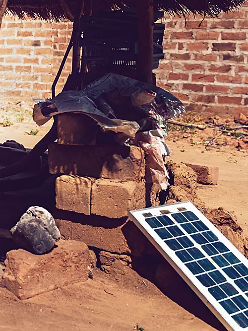 An African woman cooks in an open-air hut with solar equipment nearby.