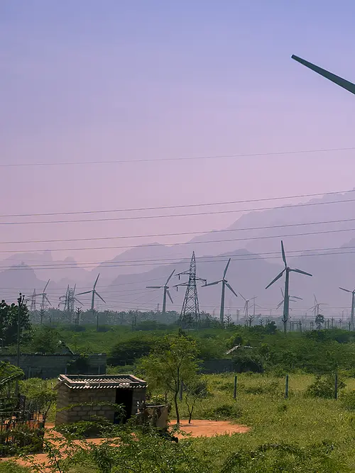 Wind turbines and power lines above lush green trees with mountains in the background.