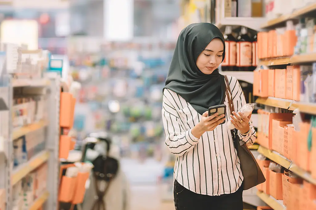 A Malaysian woman in a head scarf checks her smartphone while shopping in a pharmacy.