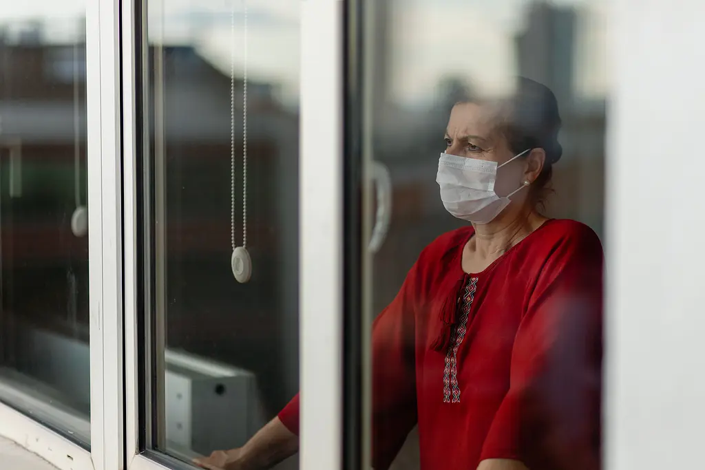 A senior woman wearing a surgical mask looks out of her window.