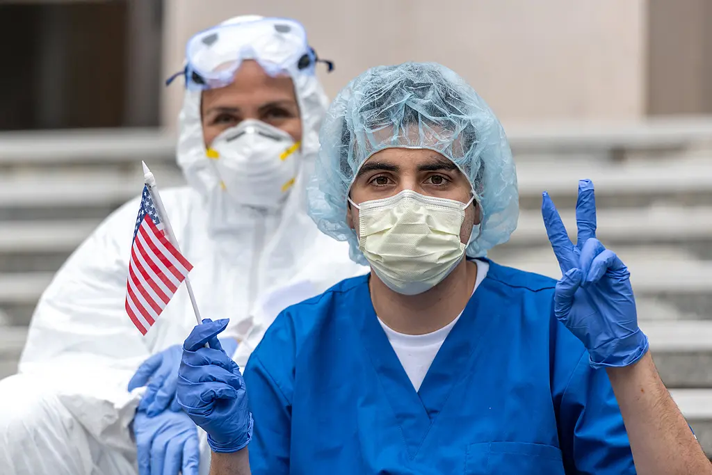 A male health worker in protective gear holds an American flag and flashes the "V for Victory" hand sign.