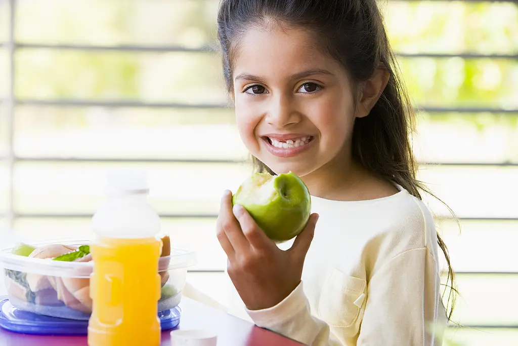 A kindergarten-age girl smiles while eating her lunch.
