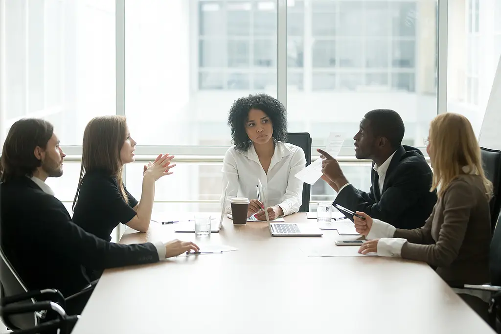 A multiethnic group of people in business attire gather around a conference table for a serious discussion.