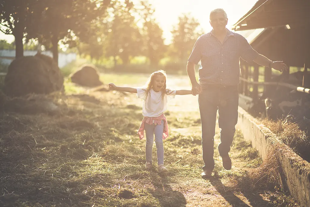 A smiling granddaughter and grandfather walk near a barn on a farm.