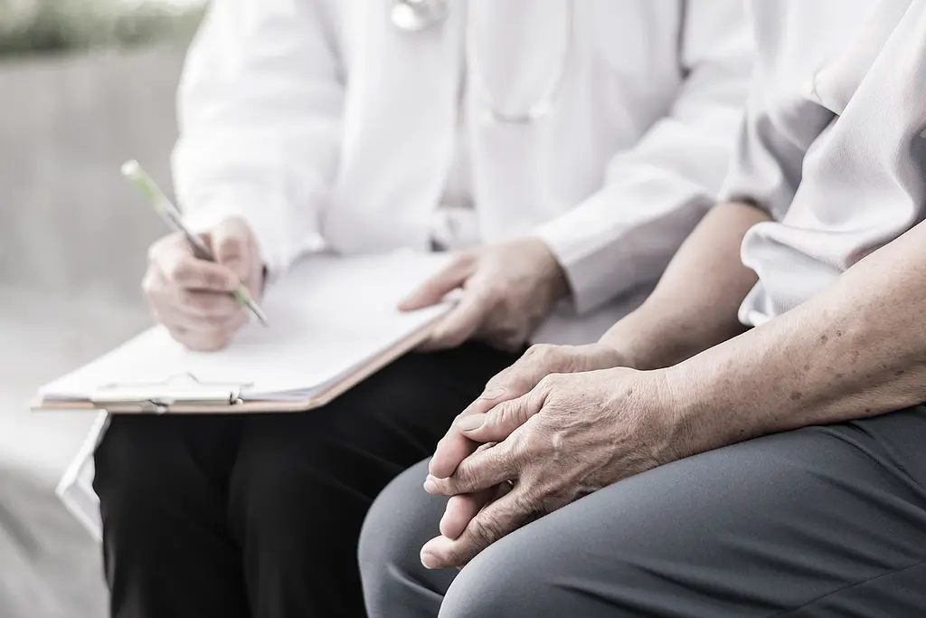 A medical professional in a white coat takes notes on a clipboard while talking to a senior patient.