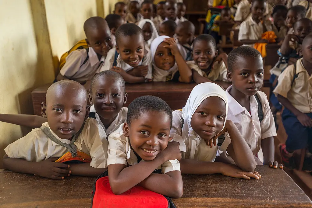 African schoolchildren crowd onto benches in a classroom and smile for the camera.