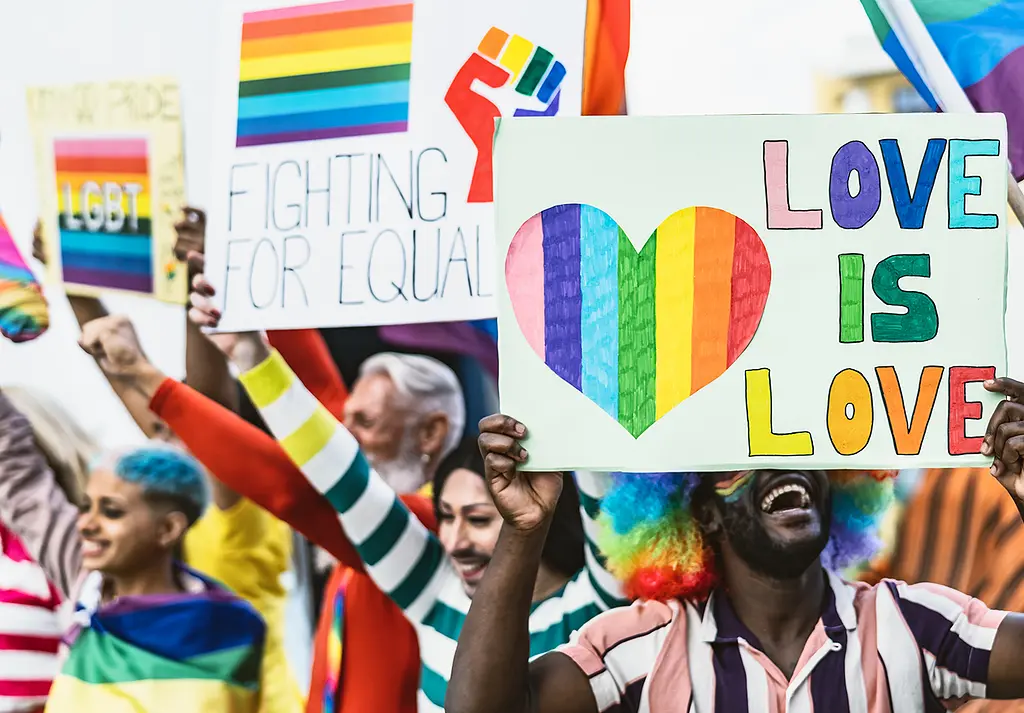 People attend a Pride parade wearing rainbow clothing and carrying "Love is Love" signs.