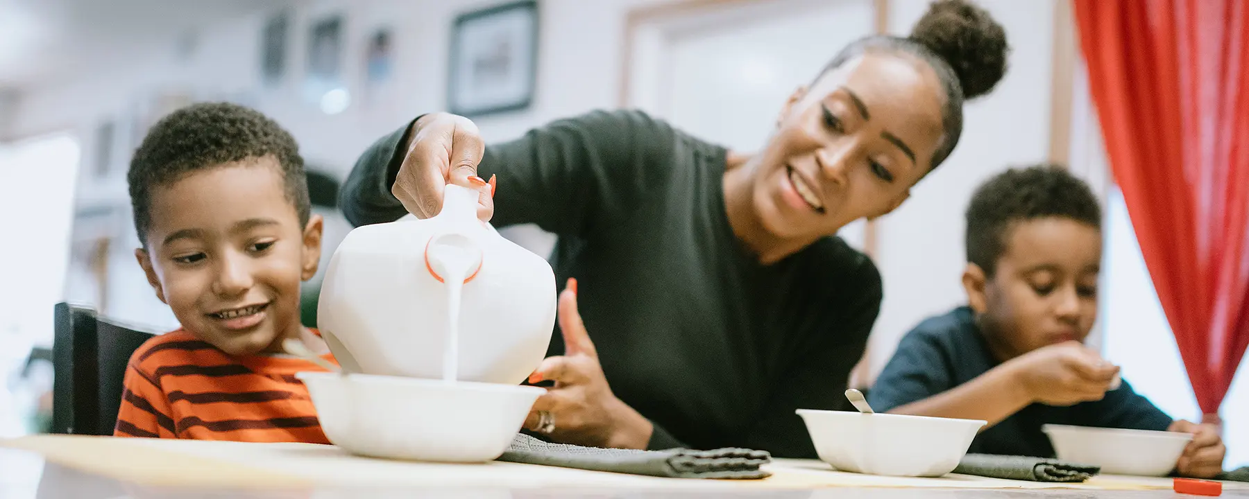 A mother pours milk into a bowl as she sits at a kitchen table with her sons