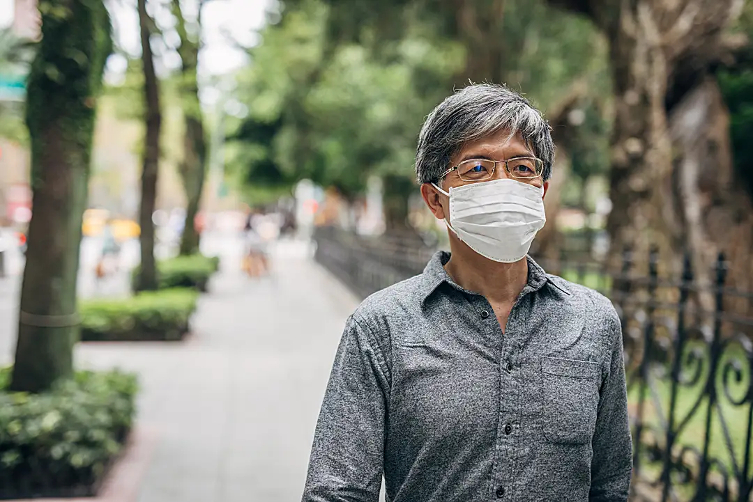 An Asian senior man wears a face mask and walks along a tree-lined sidewalk.