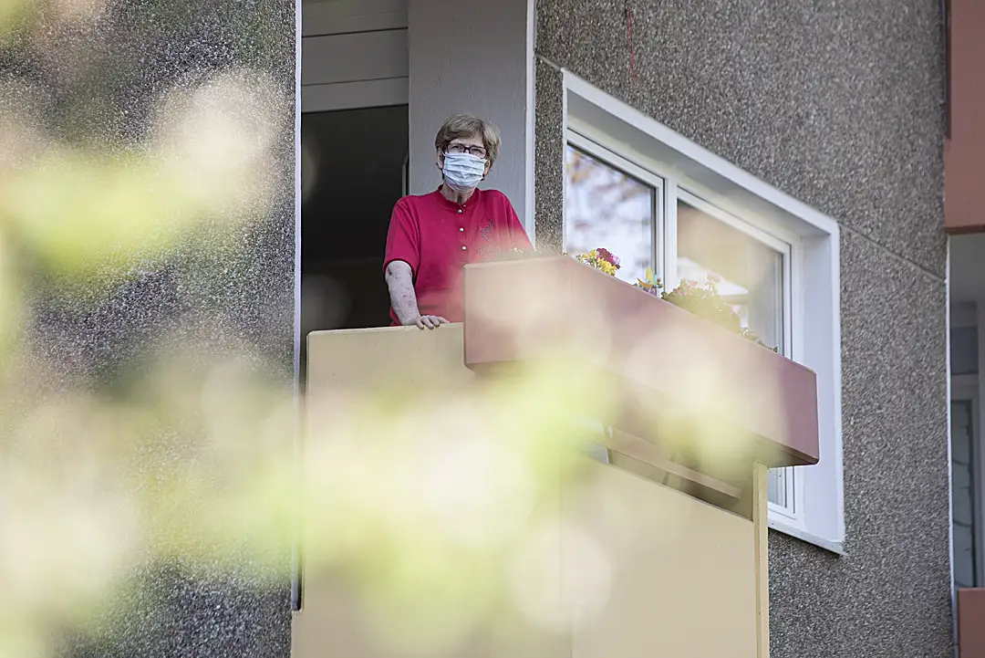 A senior woman wearing a mask looks out from a balcony.