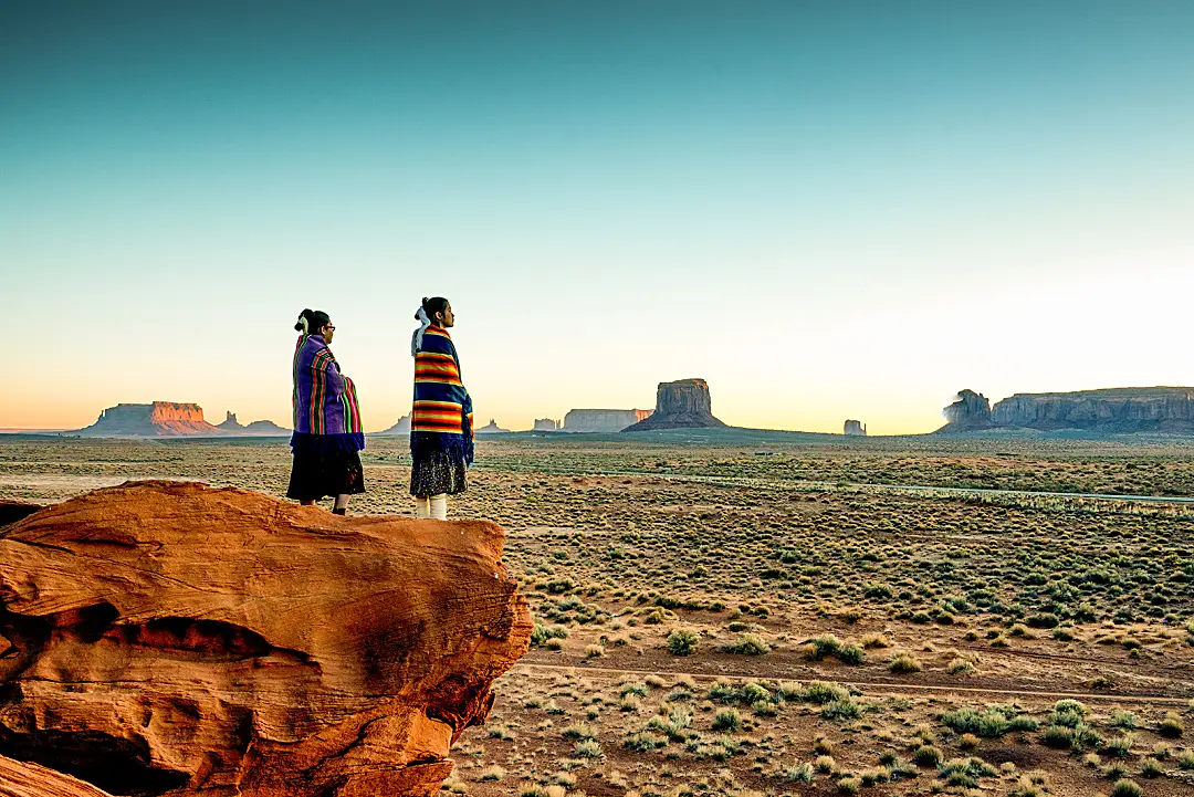 Two Navajo sisters in traditional clothing watch a sunset in Monument Valley.