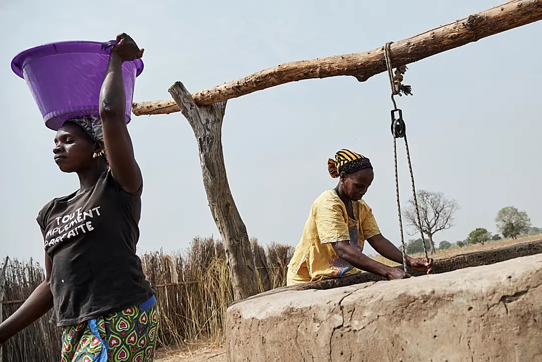 Photo of two African women working outside near a well. One is carrying a purple bucket on her head.