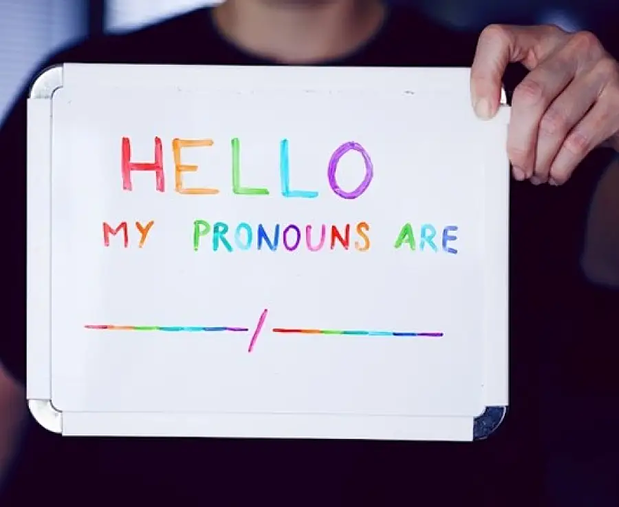 Photo of someone holding a whiteboard containing the words "Hello, my pronouns are" in rainbow colors