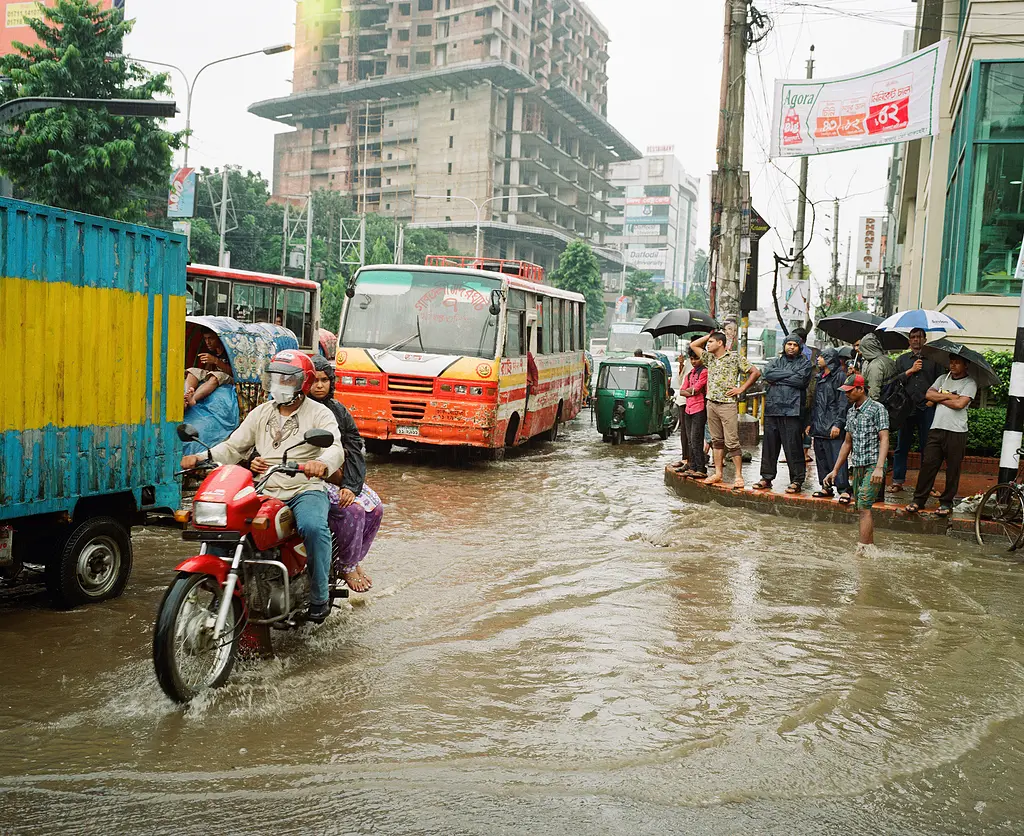 Flooding in Dhaka. People standing on the sidewalk. A motorcycle riding through the flooding.