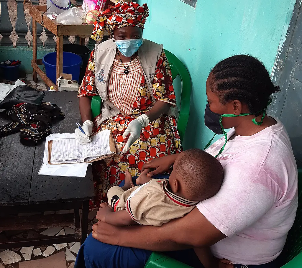 An African health worker wearing a surgical mask talks with a mother and a young boy.