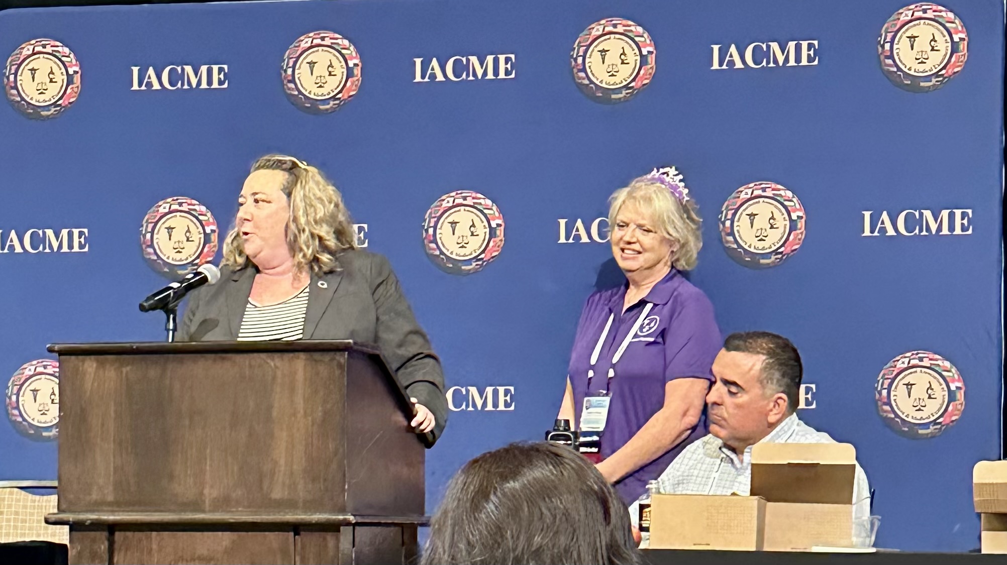 A woman speaks at a podium while two attendees listen and smile in a conference setting.